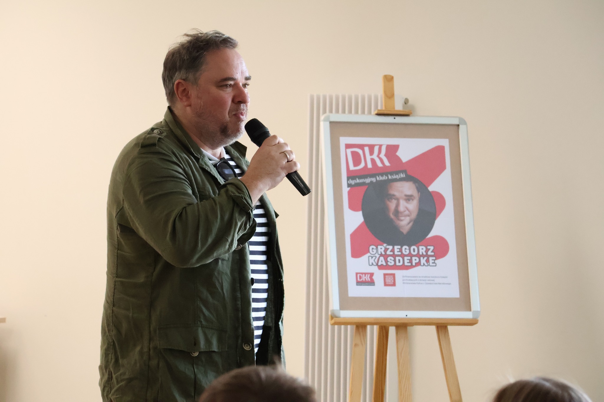 Fot. BP w Mońkach. Autor mówi przez mikrofon, za nim na sztaludze plakat promujący spotkanie autorskie z logo DKK