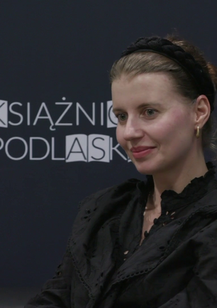 Joanna Glogaza siedzi na tle baneru z logotypem Książnicy Podlaskiej.