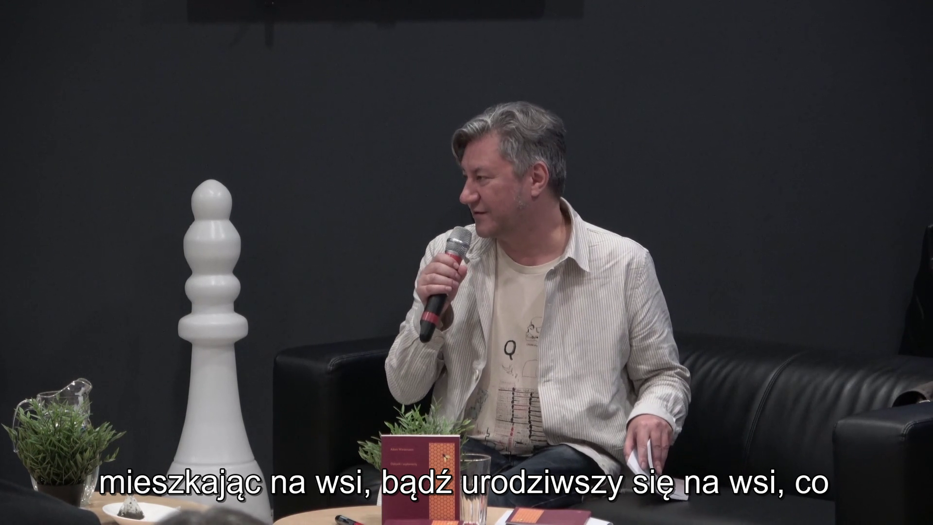 Adam Wiedemann siedzi na kanapie czarnej, trzymając mikrofon, po lewej stronie duży, biały pion do gry w szachy i mały kwiatek.