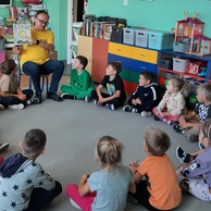 Dzieci siedzą na ziemi w bibliotece, słuchając bibliotekarza czytającego książkę.