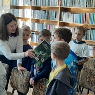 Bibliotekarka pasuje dzieci na bibliotekarzy dotykając ich ramion książką.