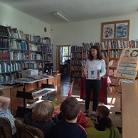 Dzieci odpowiadają na pytania bibliotekarki w sali bibliotecznej, bibliotekarka stoi i trzyma książkę, dzieci siedzą na krzesełkach.