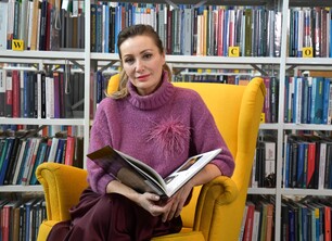 Dyrektor Iwona Gaweł Kobieta siedząca na żółtym fotelu w bibliotece, ubrana w fioletowy sweter z ozdobnym elementem. Trzyma otwartą książkę i patrzy w stronę obiektywu. W tle widoczne są półki z książkami.