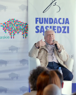 Andrzej Sapkowski siedzi w białym fotelu podczas publicznego wystąpienia. Jest ubrany w jasną, beżową kurtkę rozpiętą nad koszulą w paski oraz ciemne spodnie, a w dłoni trzyma mikrofon, gestykulując drugą ręką w trakcie mówienia. Za jego plecami widoczne są dwa banery promocyjne: jeden z kolorową grafiką żubra i napisem „Podlaskie”, a drugi z dużą, niebieską nazwą „FUNDACJA SĄSIEDZI”. Mężczyzna znajduje się na podwyższeniu, a na pierwszym planie, u dołu kadru, widać rozmazane sylwetki osób z publiczności obserwujących spotkanie.
