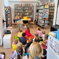 Zdjęcie nr 1 przedstawia bibliotekarkę w niebieskiej bluzce, która czyta siedzącym przed nią dzieciom książkę. Za jej plecami są regały z książkami.