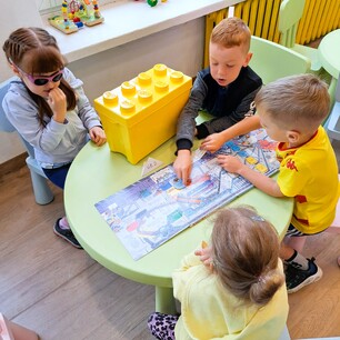Zdjęcie nr 3 przedstawia dzieci siedzące przy okrągłym zielonym stoliku i oglądające książkę o recyklingu. Na stoliku jest też żółte pudełko w kształcie klocka LEGO.