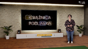 Kobieta stoi w wirtualnym studiu telewizyjnym przed dużym ekranem. Ekran wyświetla podświetlony neonowy napis KSIĄŻNICA PODLASKA. Kobieta ma na sobie czarną bluzkę w różowo-fioletowe kwiaty i niebieskie spodnie dżinsowe. Stoi po prawej stronie ekranu, trzymając ręce swobodnie przed sobą. Pod ekranem znajduje się szafka RTV z głośnikami po bokach, a na niej widoczna jest plakietka z napisem MARTA KOZŁOWSKA i pod spodem mniejszymi literami WYPOŻYCZALNIA GŁÓWNA I MIĘDZYBIBLIOTECZNA. Ściana za ekranem wygląda jak ściana z czerwonej cegły. Po obu stronach mebla RTV i ekranu znajdują się doniczkowe rośliny. U góry po prawej stronie widać logo z napisem KSIĄŻNICA PODLASKA. Podłoga jest drewniana.